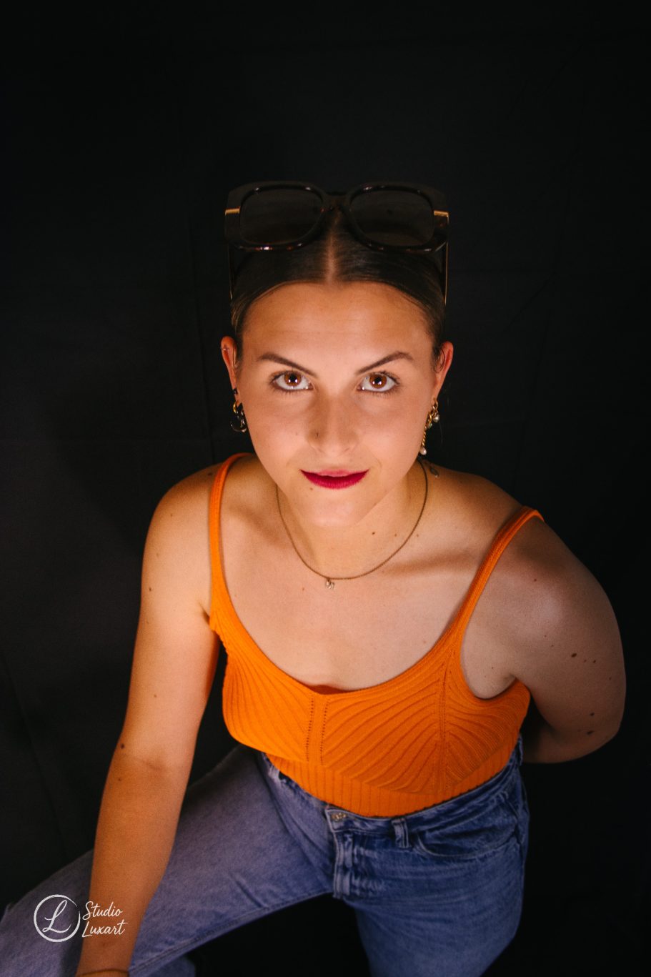 Femme souriante avec un haut orange et un pantalon en jeans, sur fond noir.