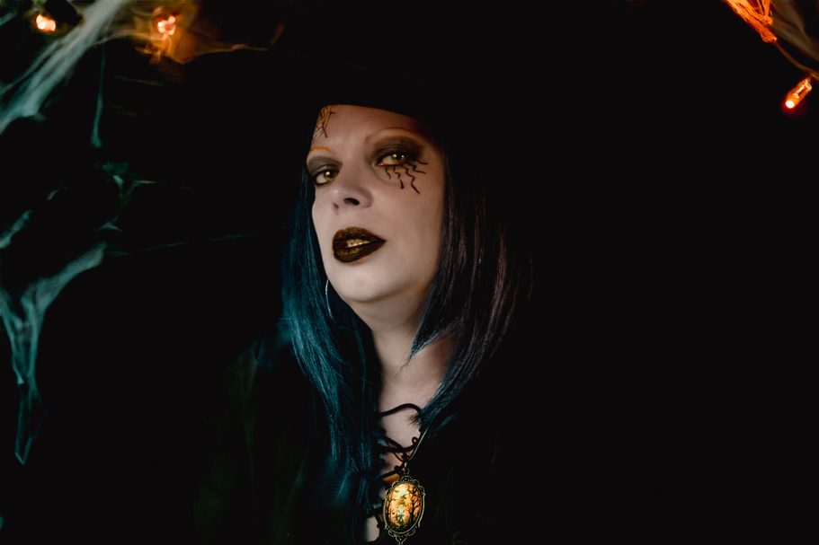Femme au visage pâle, maquillage sombre, cheveux bleus, portant un chapeau de sorcière.