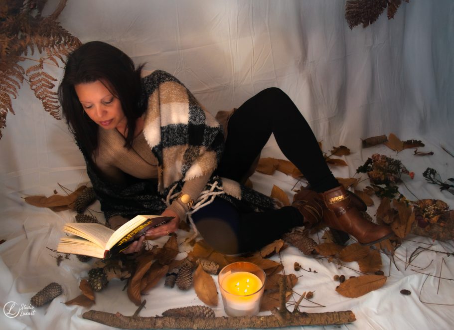 Femme allongée sur des feuilles, lisant un livre avec une bougie à côté.