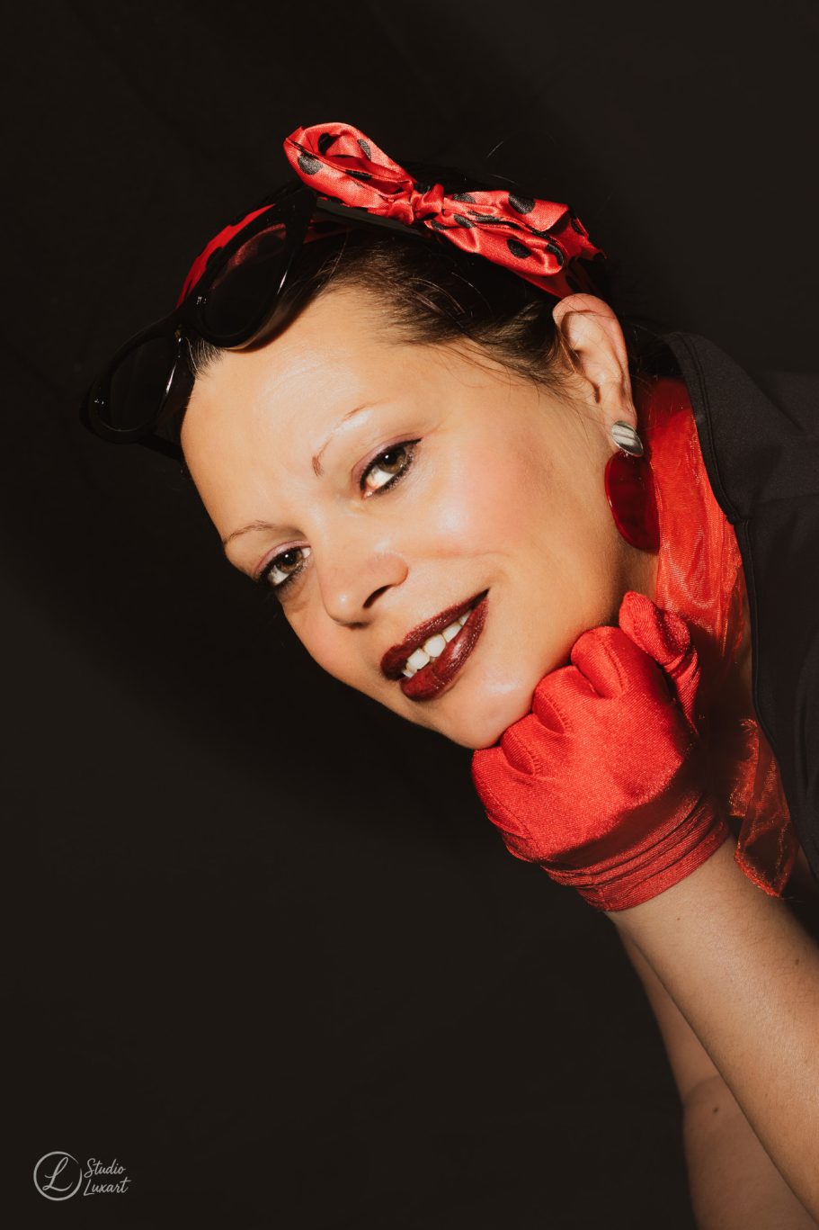 Femme au maquillage prononcé, portant un nœud rouge et des gants, souriante.