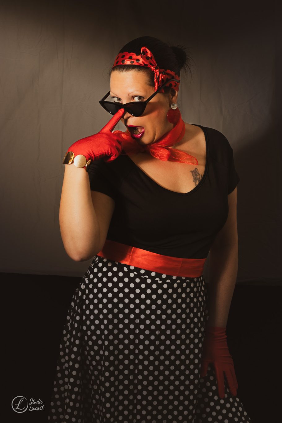 Femme en tenue rétro avec lunettes de soleil, gants rouges et ceinture rouge à pois.