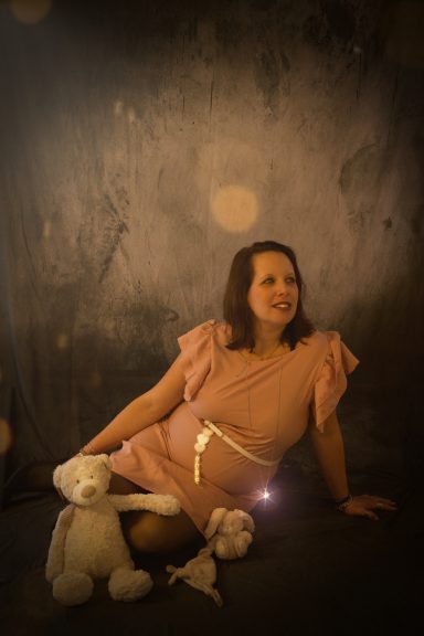 Femme assise en robe rose, tenant un animal en peluche sur fond flou et lumineux.