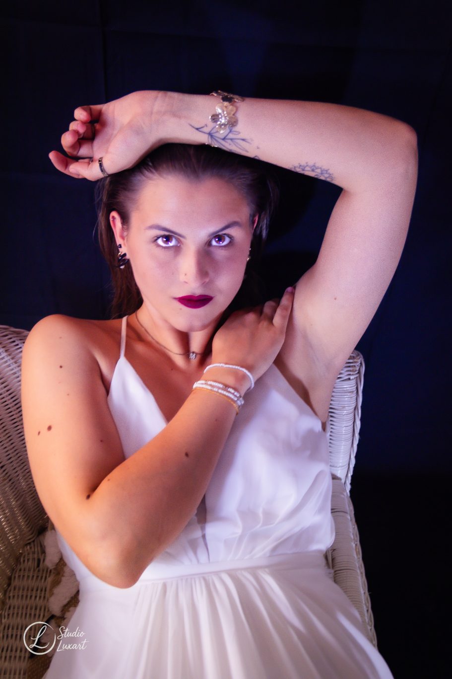 Femme dans une robe blanche, bras levé, regard intense, sur fond sombre.