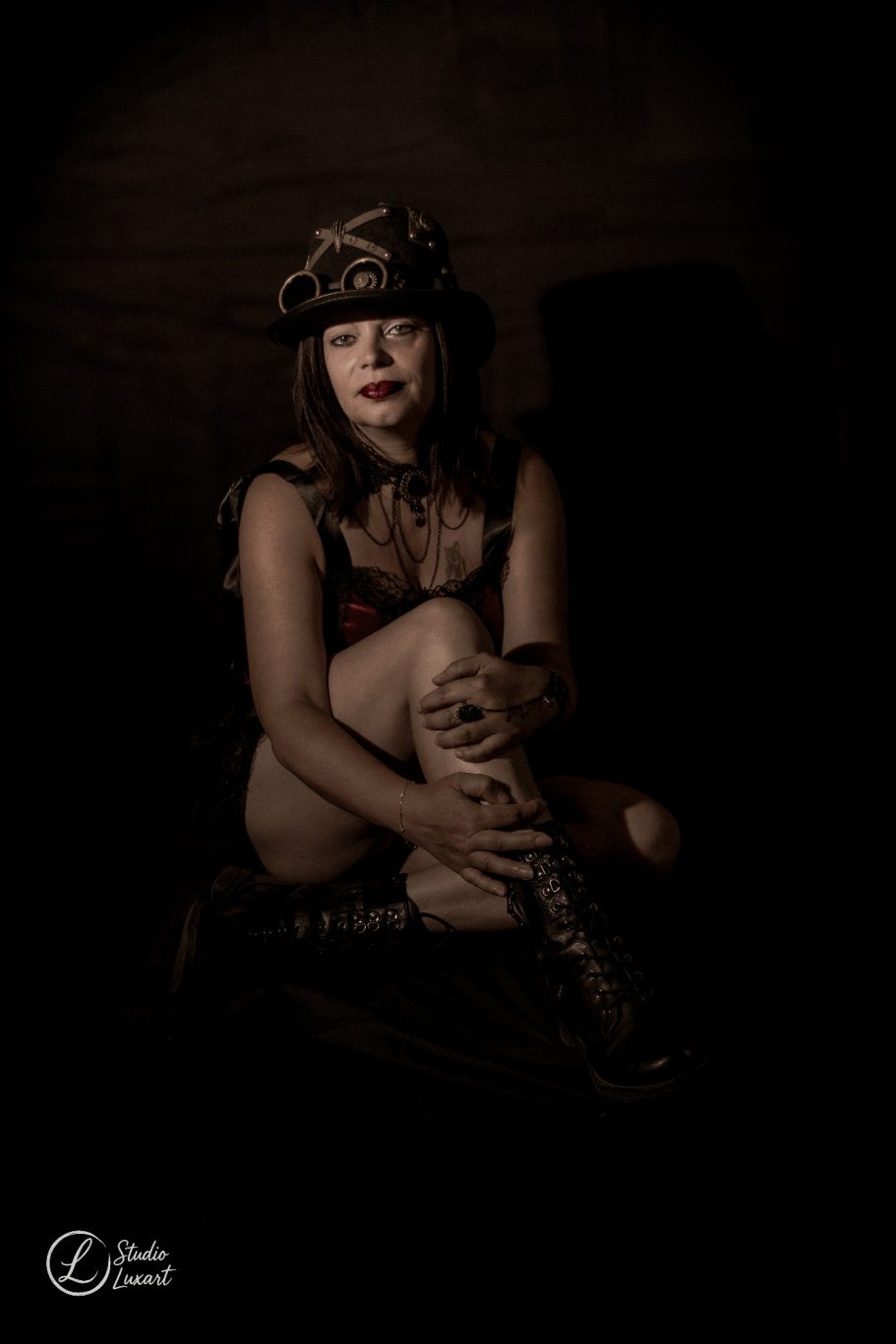 Femme assise, vêtue d'une lingerie noire, avec un chapeau, dans une lumière tamisée.