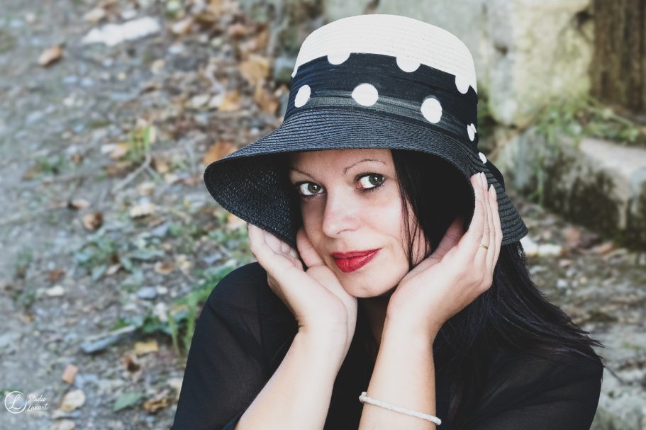 Femme avec un chapeau noir et blanc, posant avec les mains sur le visage, souriante.