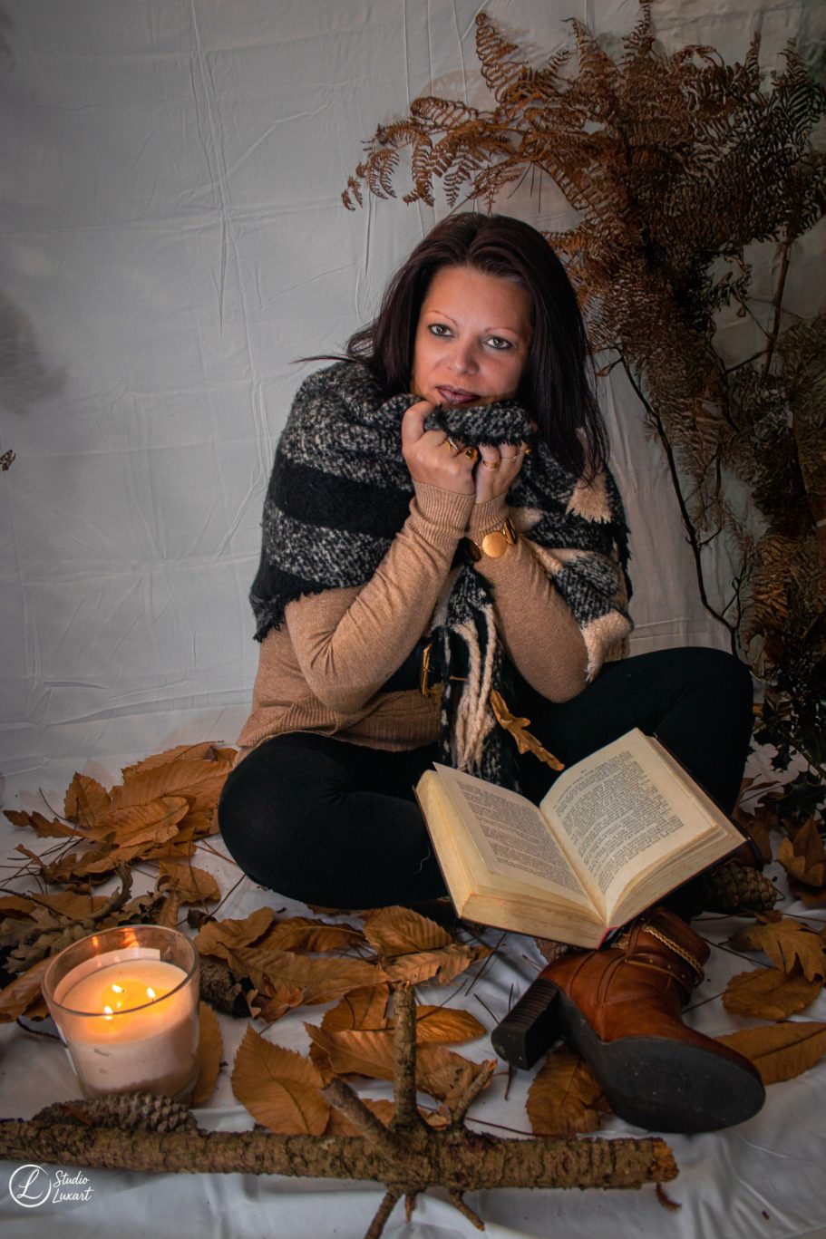 Femme assise sur un tapis de feuilles, tenant un livre, entourée de bougies et de plantes.